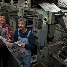 Brothers John and Art Cullen are two white men standing in a printing press, reviewing a hot-off-the-presses copy of 'The Storm Lake Times.' Image from Beth Levison and Jerry Risius’ ‘Storm Lake.’ Courtesy of 'Independent Lens'/PBS. 