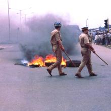 Tires burning in the middle of the road, two Indian police officers are walking by with batons in hand and crowd watching on the side of the road