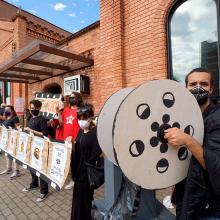 Cinemateca workers protest outside the Cinemateca Brasileira. They’re holding signs and cardboard cutouts of film reels. Photo by Benedito Faga / Alamy Stock Photo.