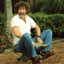 Bob Ross is white artist with a brown afro and a mostly grey beard. He is riding a bouncy alligator ride in a playground. He is wearing a white shirt and blue jeans. Image from Joshua Rofé’s ‘Bob Ross: Happy Accidents, Betrayal & Greed.’ Courtesy of Netflix. 