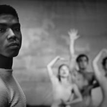 Black-and-white image of a young Alvin Ailey. He is a Black man with short hair wearing white t-shirt. Behind him are three dancers. Image from Jamila Wignot’s “Ailey”. Courtesy of NEON.