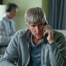 Actor Matthew Modine portrays William ‘Rick’ Singer, a white man in a grey-striped polo shirt, in ‘Operation Varsity Blues,’ a docudrama that was publicized as a documentary. Modine has grey hair and is talking on a cellphone while sitting. Courtesy of Netflix