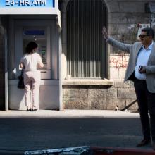 Musa Hadid, the mayor of Ramallah is a middle-aged man with a moustache. He is waving to someone and is wearing sunglasses, and is wearing a grey jacket with a shirt and trousers. There are passersby and an ATM machine behind him. Image from David Osit’s 'Mayor.' Courtesy of David Osit and POV.