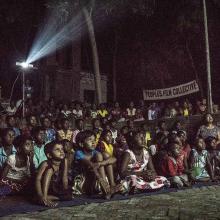 A group of Indian children and adults are gathered together outside at night, watching a film from the People's Film Collective.