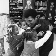 Muhammad Ali, a Black boxer, on a visit to his old grammar school in Louisville, KY, in 1977. He is surrounded by young Black children who are hugging him. From ‘Muhammad Ali,’ a four-part documentary from Ken Burns, Sarah Burns and David McMahon. Photo by Michael Gaffney. Courtesy of PBS.