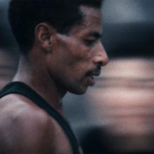 Abebe Bikila is a Black Ethiopian marathoner seen here at the 1964 Summer Olympics at Tokyo. He has black hair, a moustache, and is wearing a black vest. Image from Kon Ichikawa’s ‘Tokyo Olympiad.’ Courtesy of The Criterion Collection.