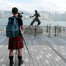 A cameraperson wearing a kilt filming a Bruce Lee statue in Hong Kong. From Mark Cousins’ ‘The Story of Film: An Odyssey’. Courtesy of Music Box Films.