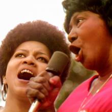 Two Black women singing a duet at the Harlem Cultural Festival. Mavis Staples (left) is wearing hoop earrings and Mahalia Jackson (right) is in a pink shirt. Courtesy of Searchlight Pictures.