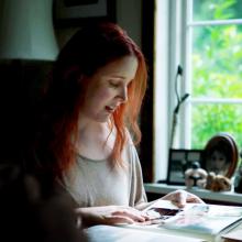 Dylan is a white woman, with red hair, in her early 30s; she is sitting at a desk next to a window, and she is looking at a photo album.