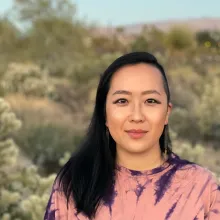 Asian-American adult is standing in front of desert landscape, wearing pink tie-dye tee, black hair passed the shoulders with left side shaved.