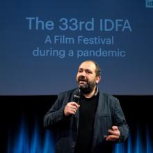The IDFA festival director, mic in hand and wearing a black jacket, addresses the audience in front of a blue curtain.