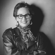 In this black-and-white portrait of Judy Irola, she has short brunette hair and glasses, she is wearing a leather jacket and she is posing with her arms crossed across her chest.