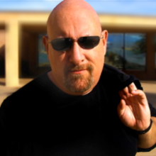 A bald man wearing black tshirt and black sunglasses.