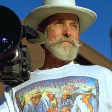 A white man with a beard and mustache wears a white hat and holds his camera on his shoulder.