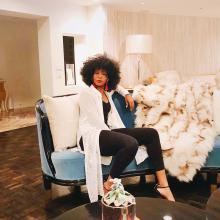  A full body shot of a Black woman in a black bodysuit and sheer white cover-up with black high heels and red dangling earrings, sitting on a couch with an afro.