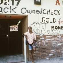 A Black woman leans against a wall that is spray painted with phrases like, 'Black owned." From 'No Loans Today.