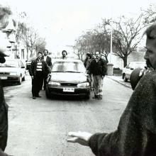 two middle-aged men are talking to each other in the middle of the street, facing a group of men surrounding a car