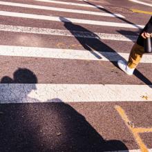 A crosswalk shows a shadow of a man using an electric wheelchair as a person walks ahead. Photo courtesy of Reid Davenport. 