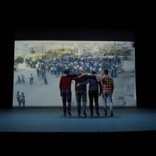 Four men doing the Dabkha dance in front of a screen showing their footage of the Syrian revolution. From Rami Farah's 'Our Memory Belongs to Us.' Courtesy of Hot Docs.