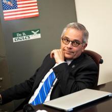 Philly DA Larry Krasner, wearing glasses, a dark suit and a blue-striped tie, sits at an office desk in front of a file cabinet.