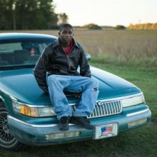 A young Black adult sitting on top of a blue car