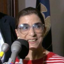 Justice Ginsburg is pictured here at a press conference, in front of a bank of microphones, and she is wearing a red-and-white striped shirt.