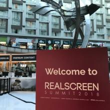 A red sign with "Welcome to REALSCREEN SUMMIT 2018" in front of an open-air business lobby with abstract metal sculpture.