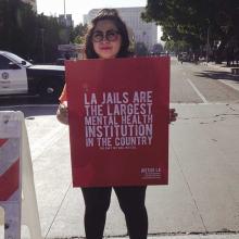 ​A woman wearing a hat and glasses, with shoulder-length hair, stands on a street and holds a sign that reads "LA Jails are the largest mental health institutions in the country." From Kenneth Paul Rosenberg’s 'Bedlam.' Courtesy of 'Independent Lens.'