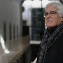 A Spanish man with white hair and glasses is staring into the distance from inside of a prison