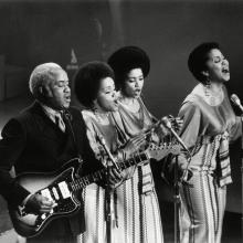 Three Black women in long skirts singing, one Black man playing the guitar. They are on stage. There are three microphones.