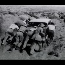A black-and-white photo of a group of people pushing a car through a barren landscape. 