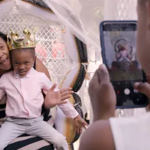 A Black boy, wearing a golden crown and a pink shirt, sits on the lap of a Black woman in a striped dress. A Black girl takes a photo.