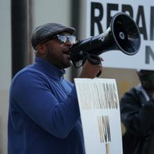 A Black man, wearing a blue polar fleece, leads a protest calling for reparations for victims of the 1921 Tulsa Massacre.