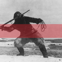 A red horizontal stripe runs through an image of a man standing on an icy landscape wielding a harpoon towards the water. Photo courtesy of ‘Nanook of the North.’
