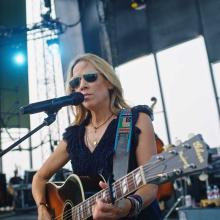 Sheryl Crow, a white female American musician with blonde hair, plays guitar and sings on a large stage. 