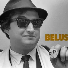 John Belushi is wearing a fedora and black sunglasses, holding up a pin with print of himself