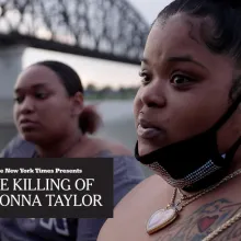 Two adult women with dark skin tone are next to a river with a bridge in the background. 