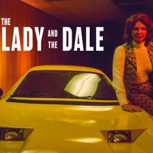 A transgender woman with brown hair to her shoulders is sitting on the side of a yellow car