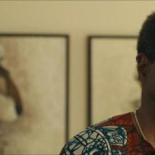 A still image from Descendant, a Black man wearing glasses and a colorful shirt standing in front of a wall inside a house.