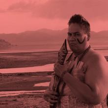 A middle-aged woman with very short hair dressed in traditional Samoan warrior clothes and holding a club near her face and standing at the edge of a lake with mountains in the distance.
