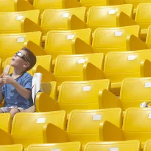 A child with white skin, short hair and wearing a blue shirt and solar viewing glasses. He sits in stadium seating eating a popsicle. 