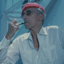 An older white man wearing a red cap, white shirt and sunglasses. He is smoking a pipe.