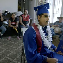 A male student in a blue graduation cap and gown and flower neckless sits among a group of people in a living room.