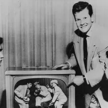 A black and white picture of several men with light skin in suits standing around a TV. 