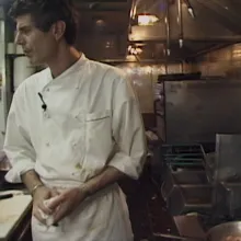 Anthony Bourdain, an older white man with short hair wearing a white chef's coat, stands in a small kitchen.