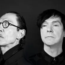 A black and white portrait of two white men both with dark hair. The man on the left has round black glasses and a pencil mustache.