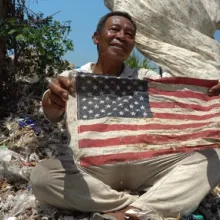 An Indonesian man holds a tattered American flag in a trash heap. 