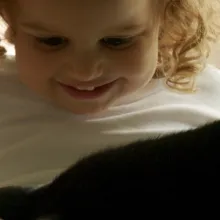 a black cat plays on the lap of a White female toddler.