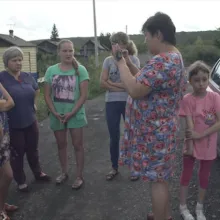 Homemaker-turned-journalist Natalia Zubkova in a blue and pink flowered dress, interviews five women with her cell phone in a rural village as her young daughter, in pink shirt and capri pants, waits nearby.