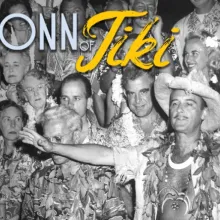 A black and white photo of a large group of middle-aged and older partygoers with "tiki" style clothing, a man in the center holds out his arms wide.